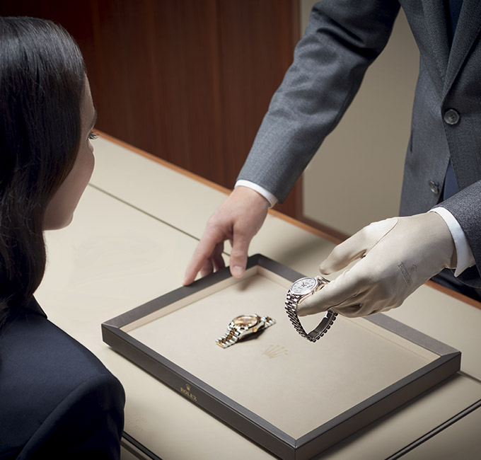 Vendedor de terno usando luva branca exibindo dois relógios Rolex para mulher